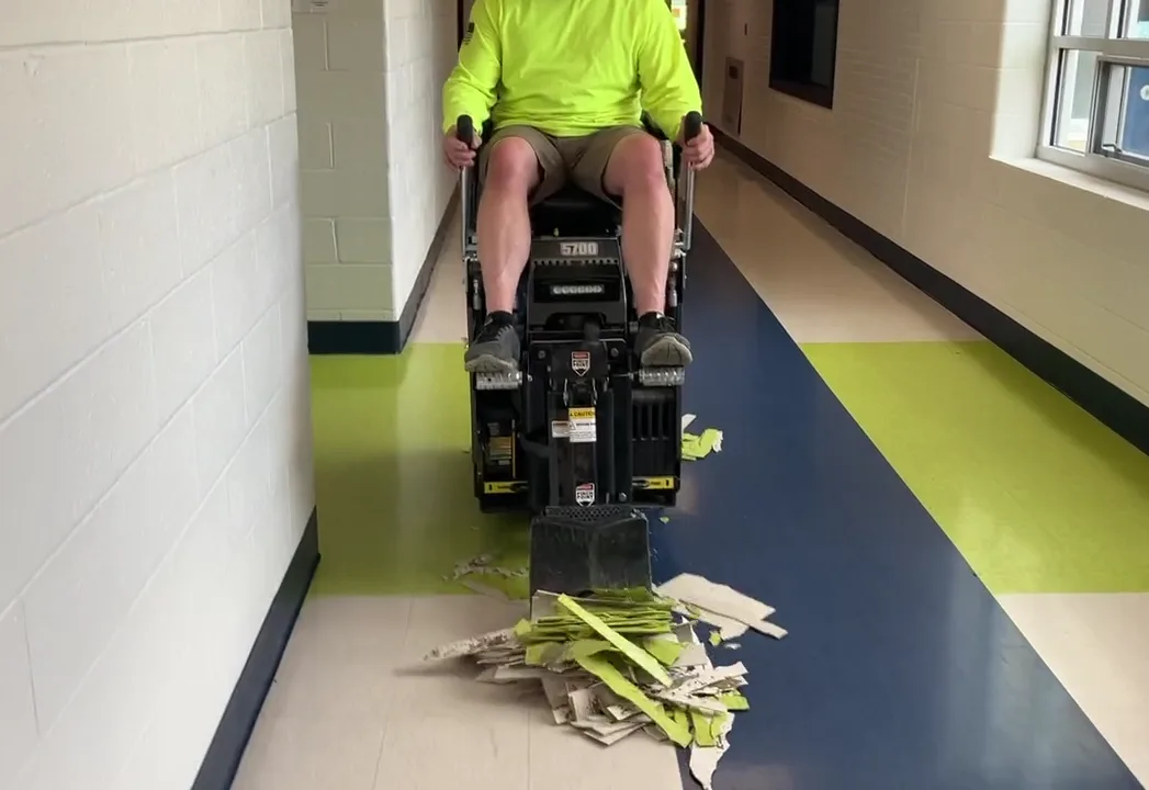 Ride-on floor scraper lifting VCT in a long hallway, the removed tiles piled ahead of the blade